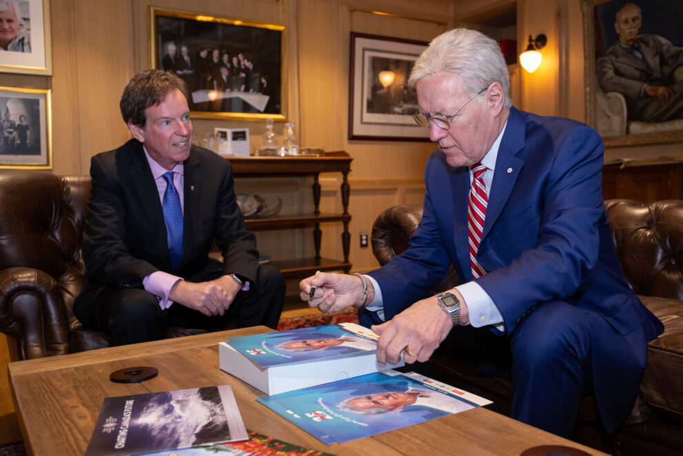 Alex Trebek signs posters while sitting across from John Geiger, RCGS CEO