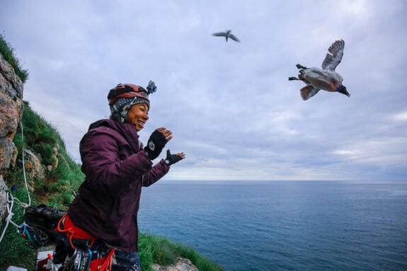 Emily Choy stands on a sea-side cliff as thick-billed murres fly overhead